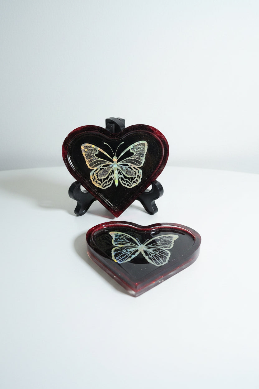 Two heart-shaped resin trays on a white surface against a light gray background. One tray is on a stand, the other is flat on the table.  Both show a holographic butterfly design on a deep red translucent heart.