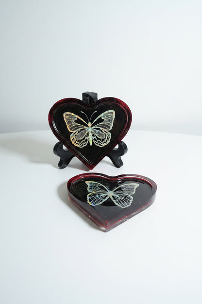 Two heart-shaped resin trays on a white surface against a light gray background. One tray is on a stand, the other is flat on the table.  Both show a holographic butterfly design on a deep red translucent heart.