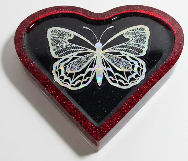 A heart-shaped resin tray with a glittery red border and a black interior, featuring a holographic butterfly design with iridescent shimmer.