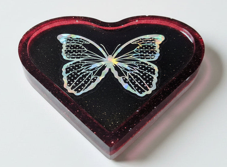 A heart-shaped resin tray with a black glittery interior and red glitter border featuring an iridescent butterfly design, set against a plain white background.