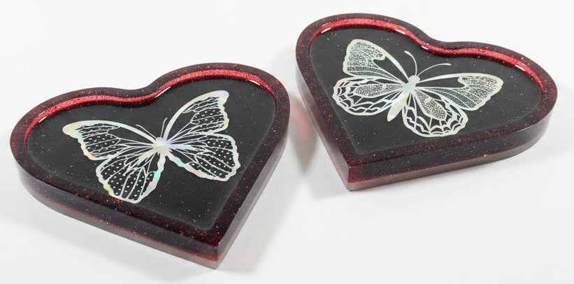 Two heart-shaped resin trays with a dark red translucent border and a holographic butterfly design in the center, displayed on a white surface.