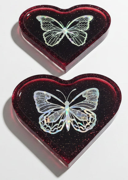 Two heart-shaped resin plaques with embedded glitter and a holographic butterfly design, displayed on a clean white background.